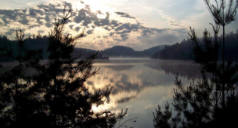 Gentle light is reflected on still water, surrounded by hills and trees.